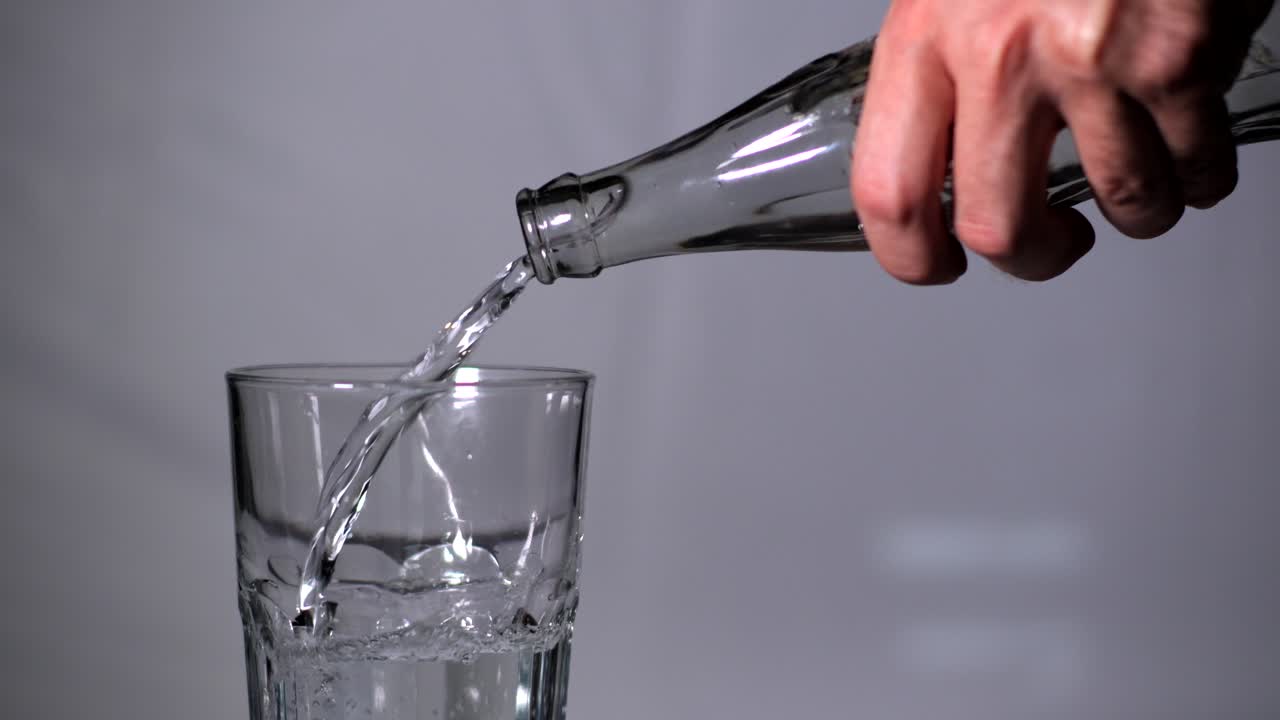Man's Hand Pouring Water Into a Drinking Transparent Glass From Bottle on Studio Background - closeup