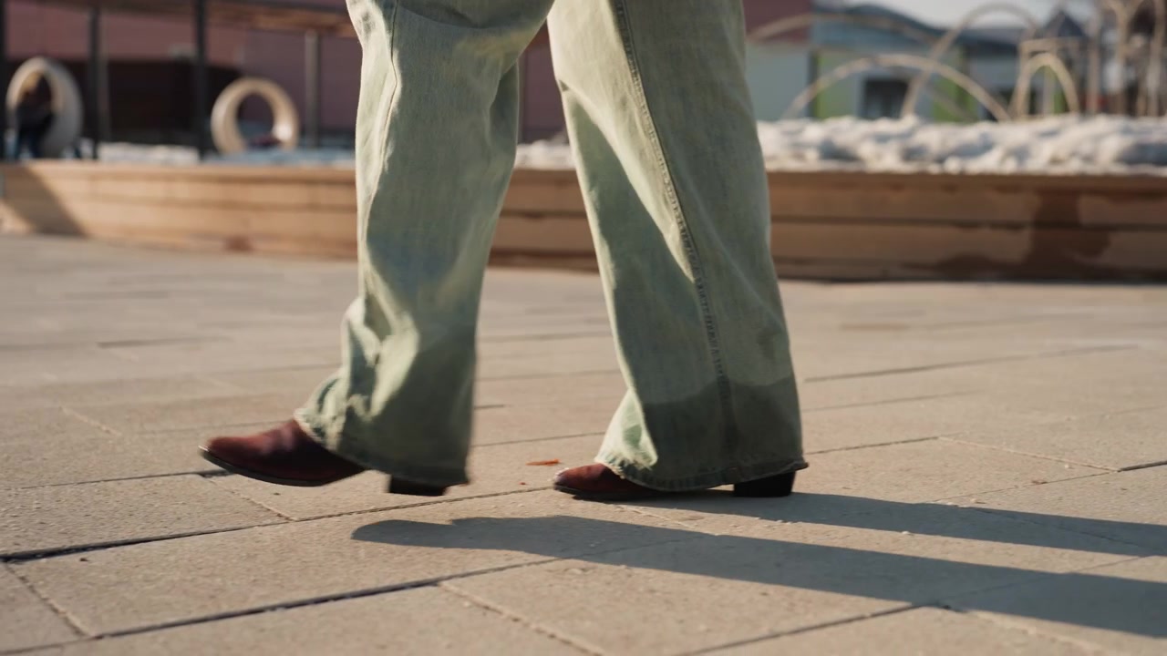 Pretty green pants walking on promenade, brown loafers gliding across sunlit tiled pavement near harbor, cropped low angle view, casual model stride, gentle shadow elongation, vintage flare hem
