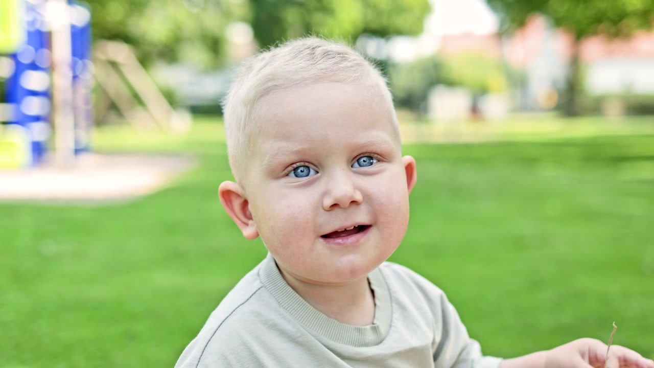 Ein Kind mit hellem Haar und blauen Augen spielt tagsüber in einem Park, während es auf Gras in der Nähe von Spielgeräten sitzt.