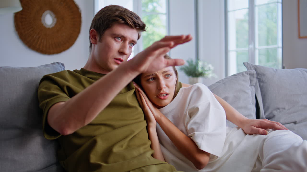 Married couple watching horror movies at cozy home. Man embracing scared woman