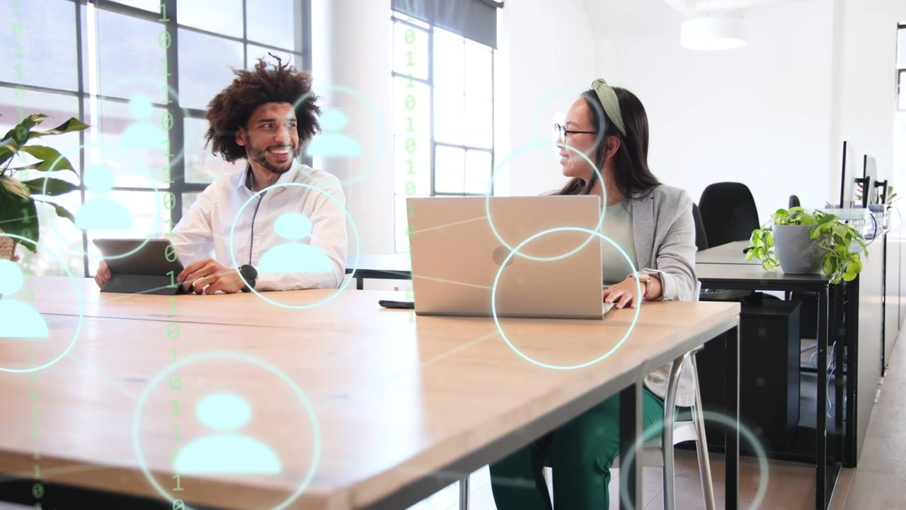 Colleagues talking and reacting in tech office, cyan user icons appearing over tablet and laptop