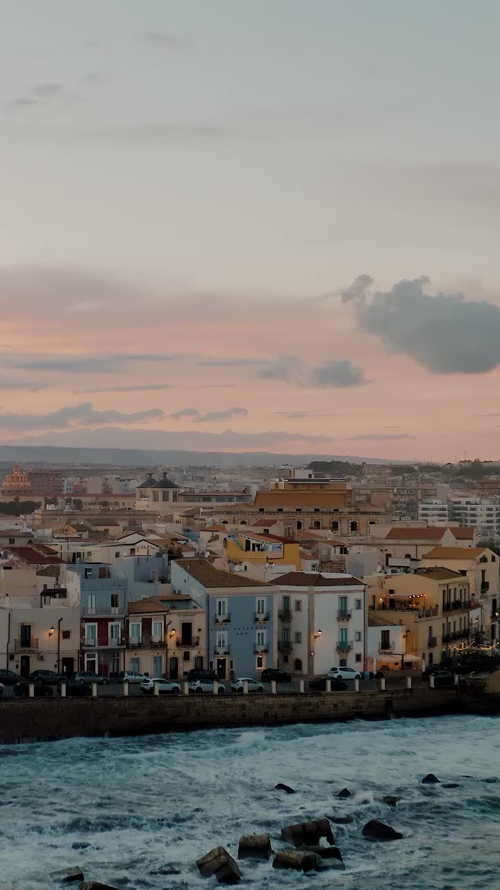 Vertical aerial view of Ortigia island in beautiful evening at sunset. Drone shot of historic Italian town in Sicily by the Mediterranean sea. Large waves crashing onto the shore with colorful sky.