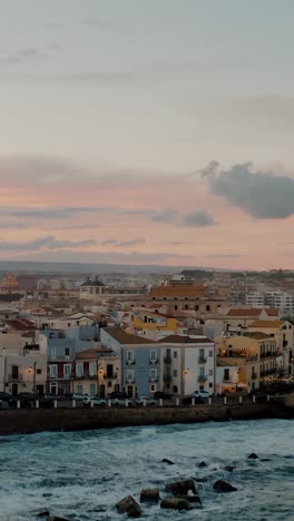 vertical aerial view of ortigia island in beautiful evening at sunset. drone shot of historic italian town in sicily by the mediterranean sea. large waves crashing onto the shore with colorful sky.