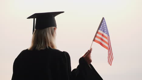 Absolvent-Im-Mantel-Mit-USA-Flagge-In-Der-Hand-Trifft-Die-Morgendämmerung