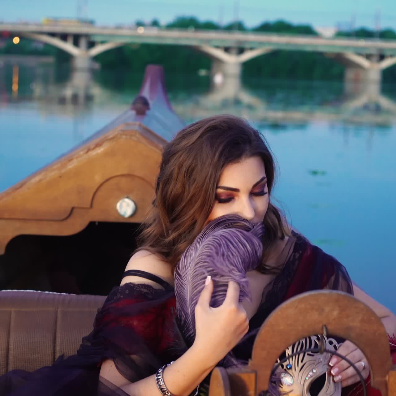 A charming woman with a bright make-up is holding a violet feather in her hand and smiling in the evening on the gondola. Blurred Background