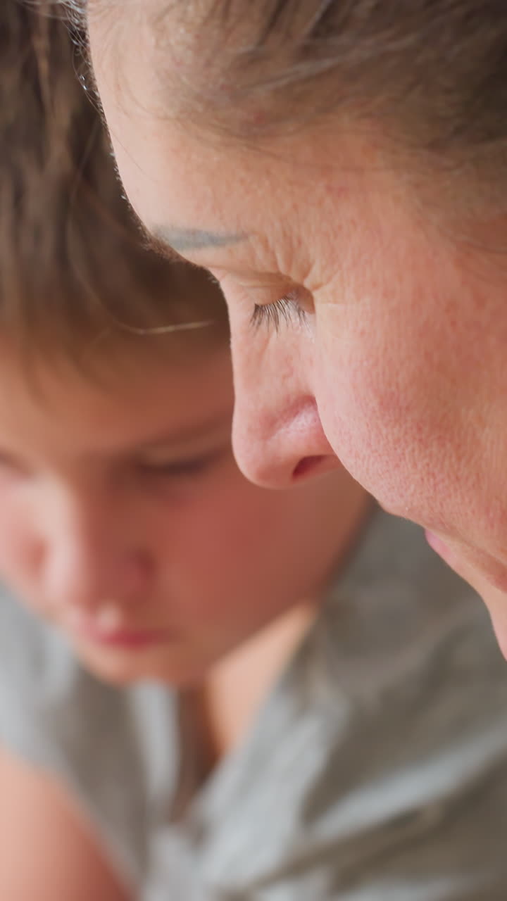 클로즈업된 흰색 배경의 엄마와 아들이 조용한 과외 시간을 보내고 있어요. 부드러운 지도와 세심한 집중으로 실내에서 학습하는 모습이에요. 아이 옆에서 엄마의 옆모습이 보이며, 애정과 인내심을 보여주고 있어요. 부드러운 초점의 인물 사진입니다.