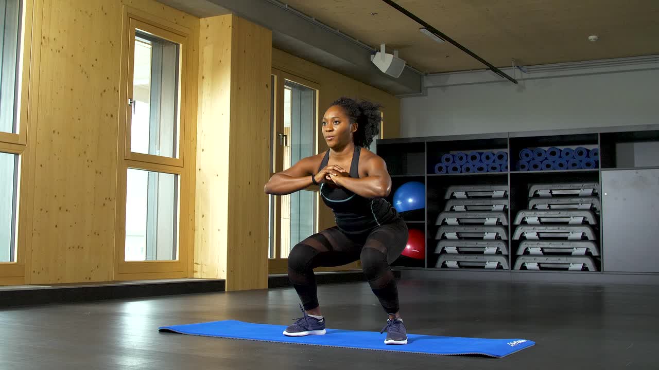 mujer africana preparándose para el entrenamiento en un gimnasio, de pie con confianza, haciendo cuclillas, en un entorno interior bien iluminado