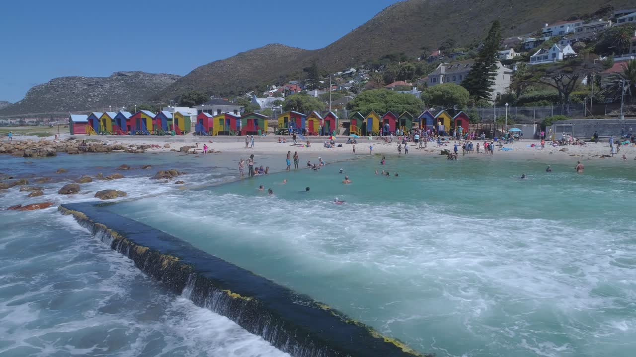 파도가 바다 조수 웅덩이에 부딪히면서 무인 항공기가 공중에 떠 있고, 배경에는 색색의 해변 판잣집이 있습니다.