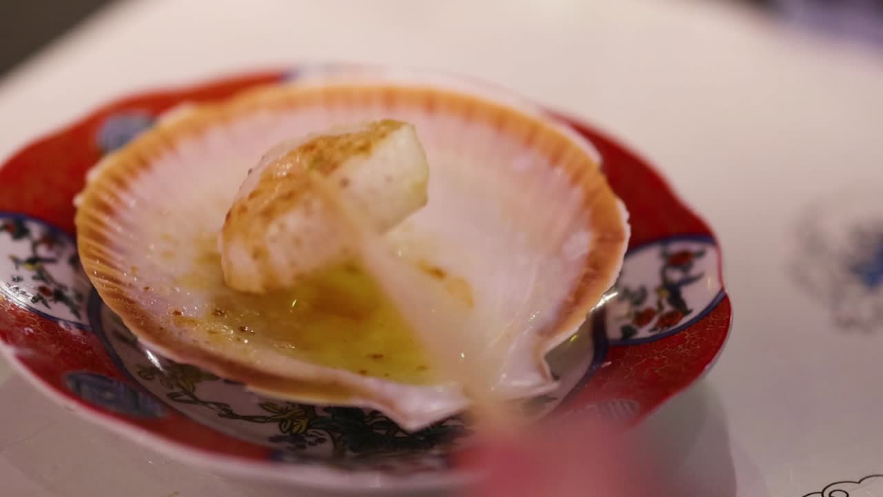 A scallop is delicately lifted with chopsticks from a decorative shell plate, showcasing culinary presentation.