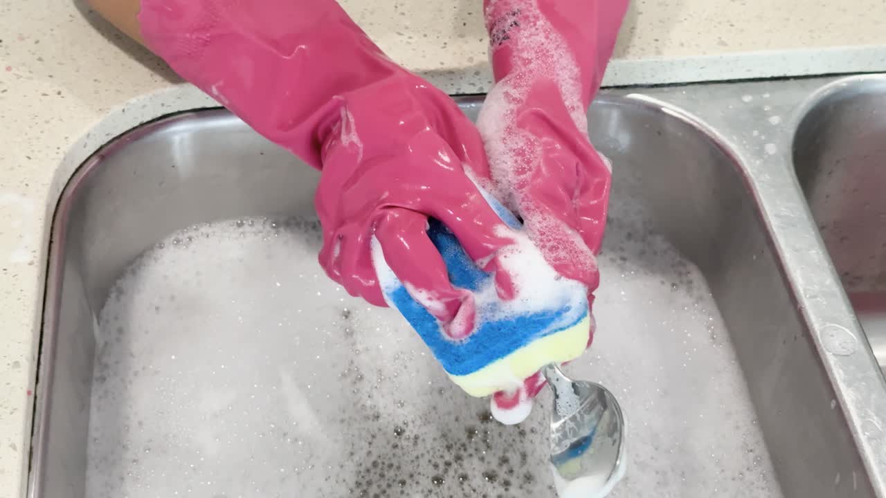 A person wearing pink gloves thoroughly scrubs dirty spoons with a blue sponge in a stainless steel kitchen sink filled with soapy water, under bright lighting