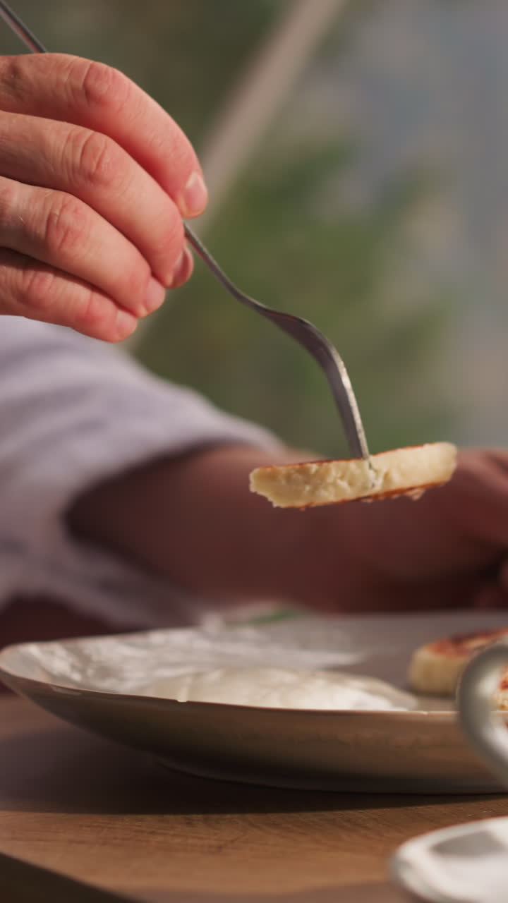 Man dips slice of pancake in sour cream. Man enjoys breakfast in glamping house against glass wall with panoramic view. Room service in glamping town