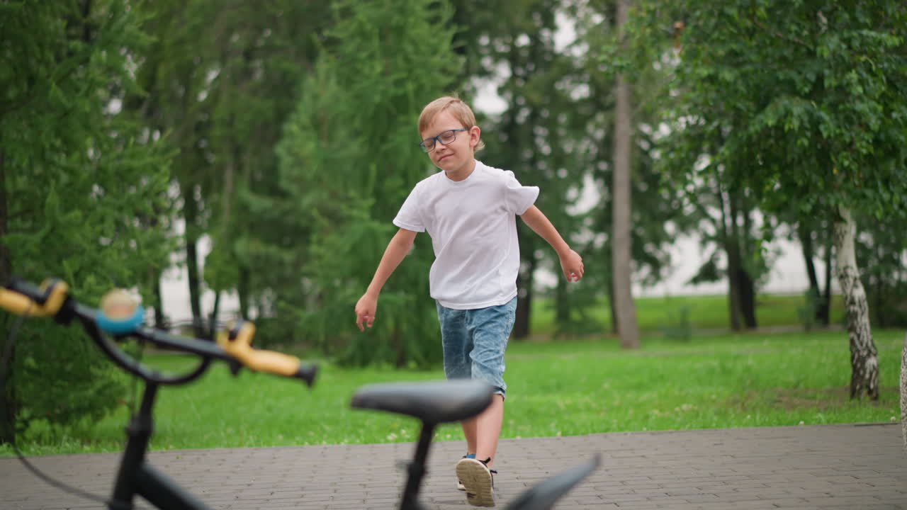 A young boy, wearing glasses, jumps joyfully toward a parked bicycle with a determined look and a stern smile on his face, dressed in a white top and denim shorts