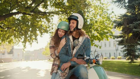 Happy-couple-brunette-girl-in-a-plaid-shirt-leans-and-hugs-her-boyfriend-with-curly-hair-in-a-White-moped-helmet-and-denim-jacket,-who-sits-on-a-moped-in-the-summer-city