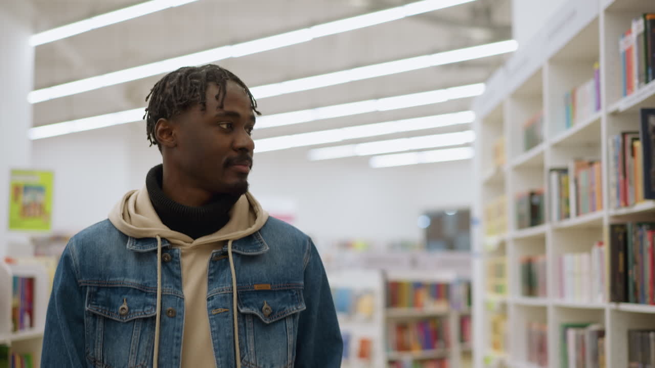 Close-up shot of man walking and browsing in store, dressed in denim jacket, hoodie, and scarf, with colorful book shelves in background and bright lighting, exuding calm and relaxed vibe