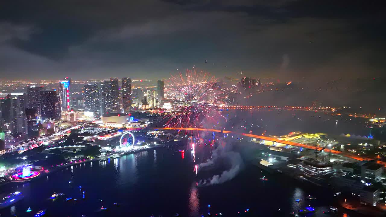 A drone hovers above a packed bay while fireworks thunder into the sky, captured in smooth cinematic motion
