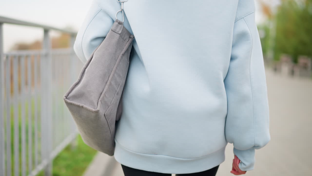 Partial view of young woman in casual wear walking with bag, with a blurred background featuring street light poles, trees, and an iron fence in a peaceful urban setting