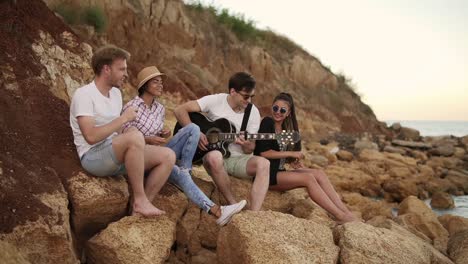 group of young hipster friends sitting on the rocks by the seashore and playing guitar, singing songs and dancing. slow motion