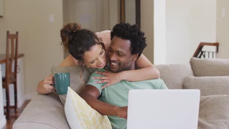Happy-biracial-woman-with-coffee-embracing-her-male-partner-sitting-on-sofa-with-laptop