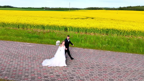 a joyful couple in wedding attire jogs down a cobblestone path next to a vibrant yellow field, blending romance with playful energy in a rural spring landscape