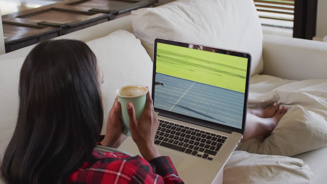 Composite of woman sitting at home holding coffee watching athletics running event on laptop