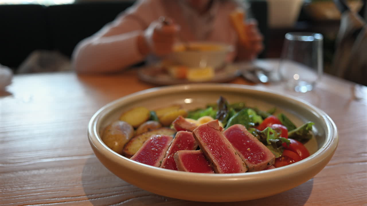 Seared tuna steak slices served with potatoes, fresh salad, tomatoes, and egg on wooden table with a woman eating in the background at a restaurant