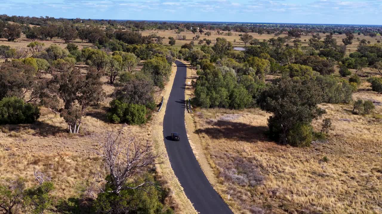 A drone follows an electric vehicle traveling along a winding road through dry, open woodland in Warrumbungle National Park under bright daylight
