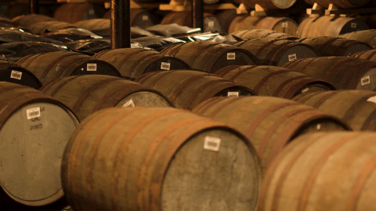 Rows of oak barrels aging whisky in warm, dimly lit distillery warehouse, slow panning shot
