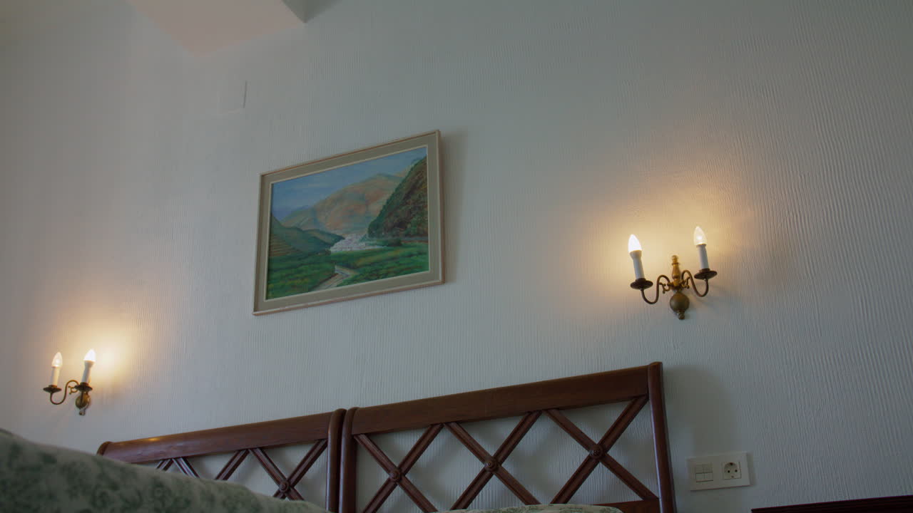 A smooth dolly shot moving from left to right inside a traditional Spanish bedroom. The camera glides past the headboard, revealing classic brass sconces and a framed landscape painting