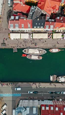 vertical top down drone shot tracking along the iconic canal of nyhavn, copenhagen, denmark. showcasing its colourful roofs, historic boats and many pedestrians walking along the waterfront.