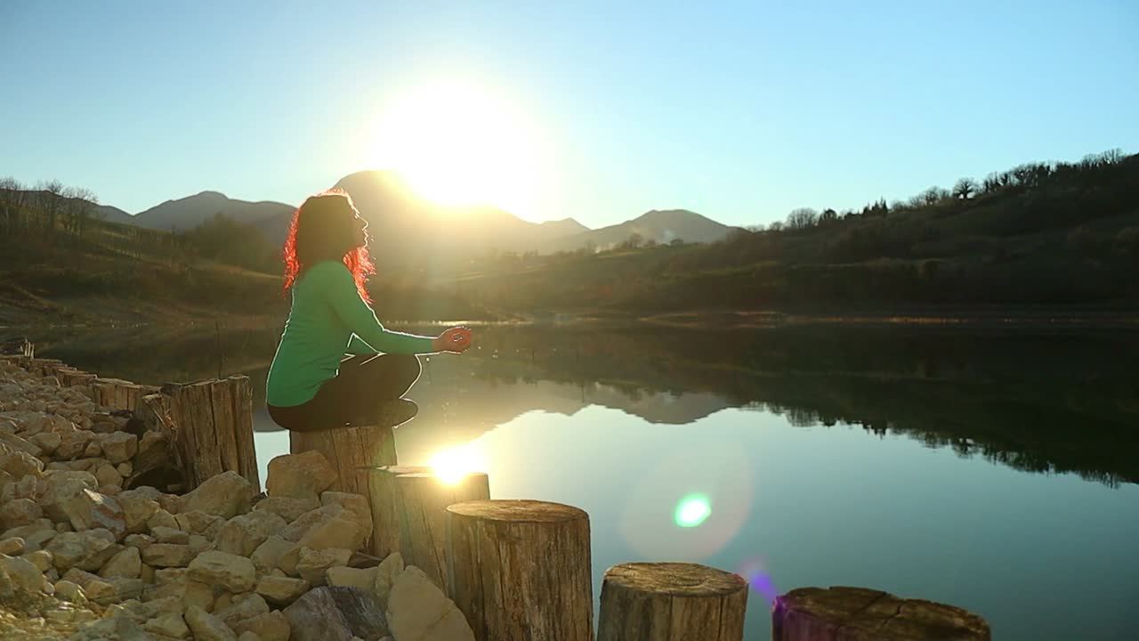 una chica caucásica hace práctica de meditación junto al lago al amanecer en un día soleado