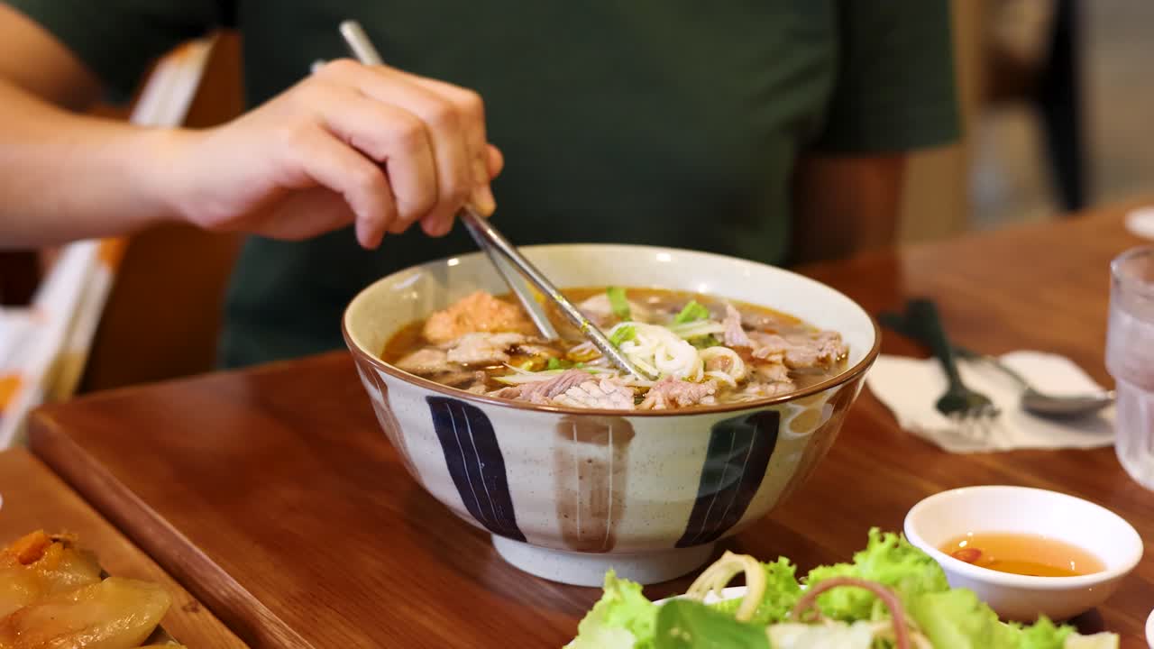 Cận cảnh đôi tay dùng đũa kim loại gắp sợi phở từ tô phở bò