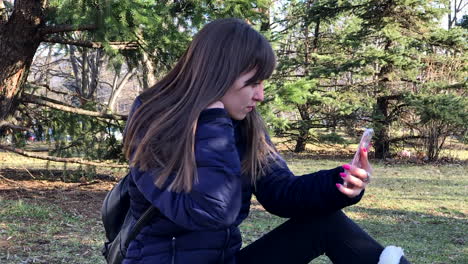 Cámara-Lenta-De-Una-Joven-Tomando-Selfies-Con-Un-Smartphone-Usando-Una-Cámara-Frontal-Sentada-En-El-Suelo-En-Un-Parque-Urbano