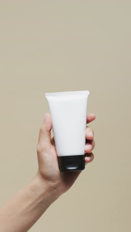 hand of woman holding tube of cream on beige background with copy space, slow motion
