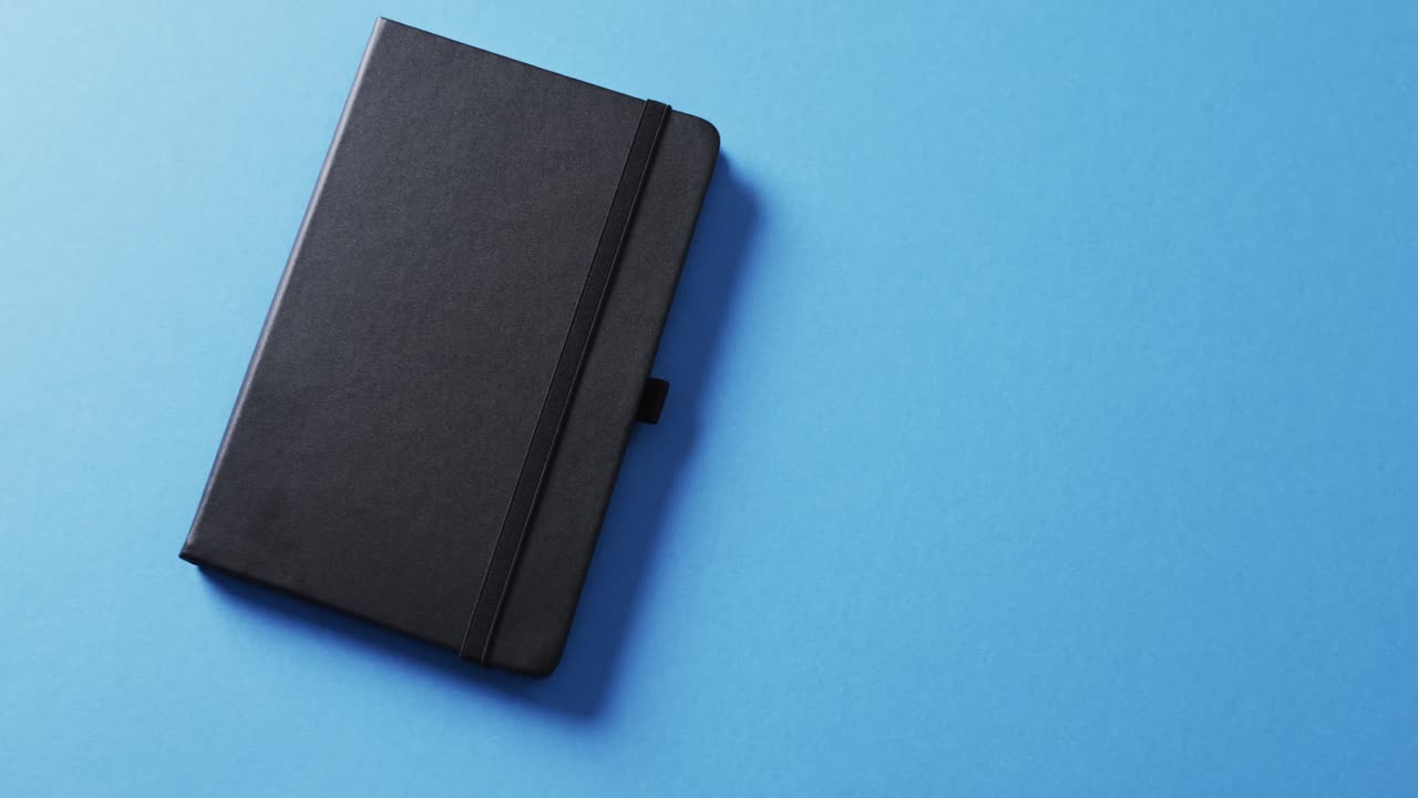 Close up of closed black notebook with copy space on blue background in slow motion