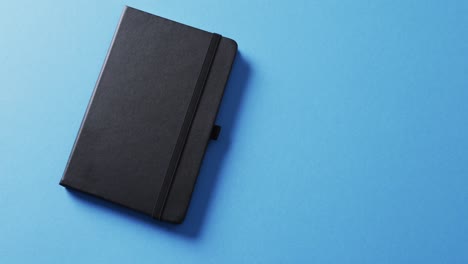 close up of closed black notebook with copy space on blue background in slow motion