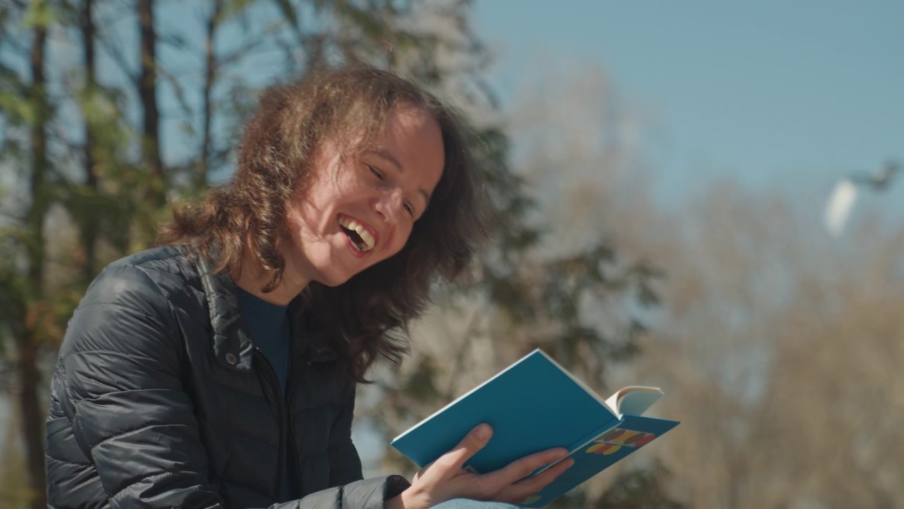 Primo piano di una donna che legge un libro blu all'aperto con alberi e cielo sullo sfondo. Il soggetto tiene un quaderno aperto mostrando un'espressione coinvolta mentre interagisce con il testo. La luce naturale soffusa illumina