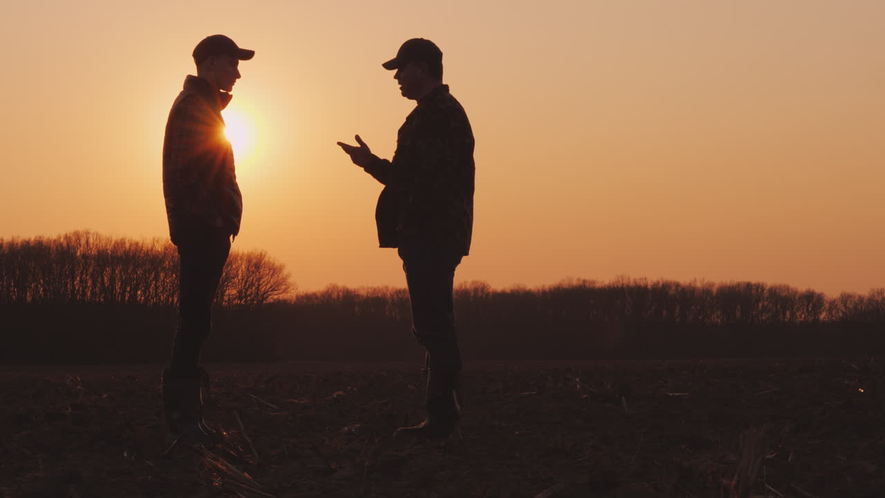 Un agricoltore anziano e uno giovane camminano insieme su un campo arato al tramonto.