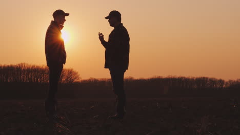 Ein-älterer-Und-Ein-Junger-Bauer-Gehen-Bei-Sonnenuntergang-Zusammen-über-Ein-Gepflügtes-Feld