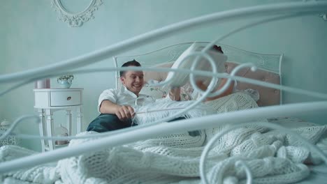 Young-beautiful-couple-having-fun-in-bed-they-fight-by-cushions-laughing-passing-camera-along-bed-slow-motion