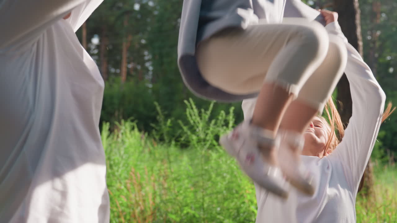 Smiling siblings enjoying sunny day on forest path, holding hands and gently lifting youngest between them, surrounded by lush greenery and warm light, sharing joyful outdoor family bonding