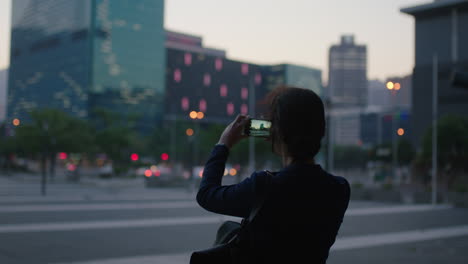 portrait of young woman taking photo of city using smartphone enjoying evening lights wearing backpack