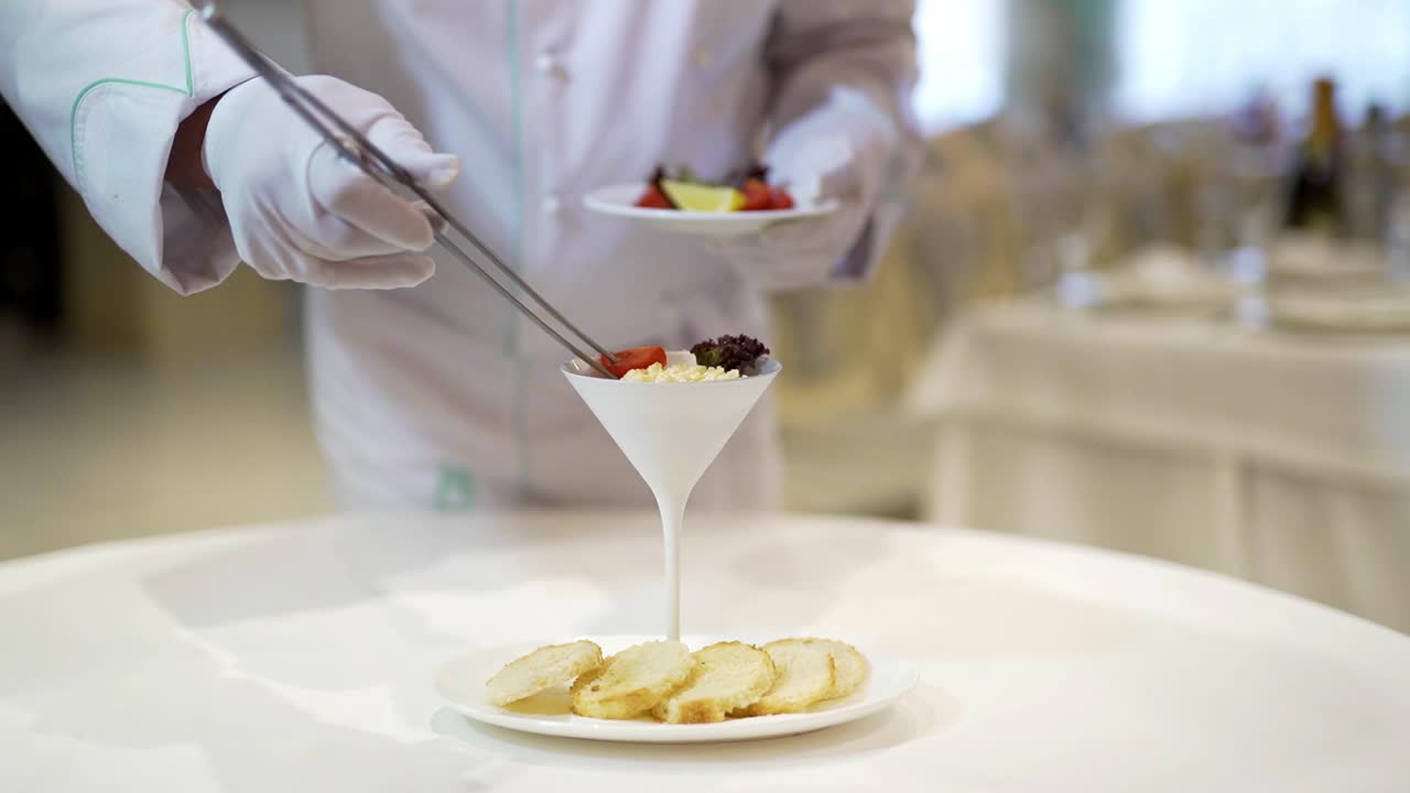 A waiter serves cottage cheese in a high ceramic bowl laying out cherry tomatoes, greens and lemon slices on the background of a white plate with five round slices of bread on the table in the restaurant.
