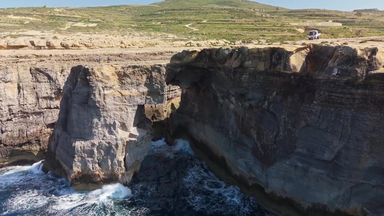 Wied il-Mielaħ natural arch in Gozo, Malta, with rugged coastal cliffs, blue Mediterranean waters and rocky shoreline, captured by drone over the scenic seaside landscape.