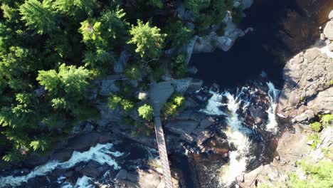 Aerial-top-view-of-Canadian-forested-cliff,-cascading-waterfall,-and-footbridge