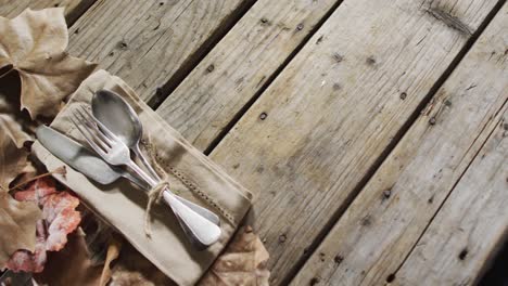 Close-up-view-of-cutlery-set-over-a-napkin-and-autumn-leaves-with-copy-space-on-wooden-surface