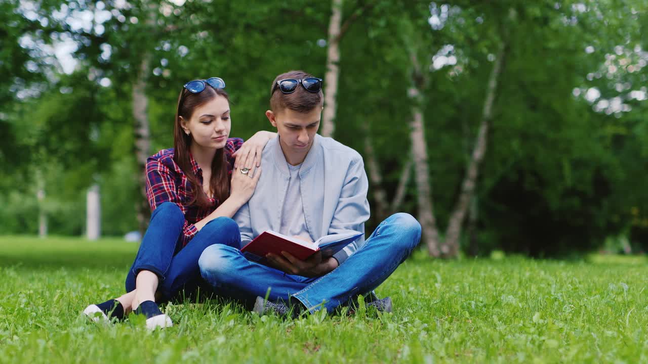 un joven y una mujer leyendo un libro en el parque se sientan en un video hd de césped verde
