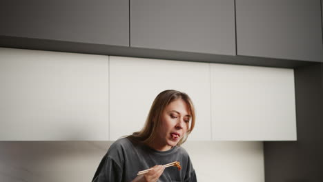 woman eating meat with chopsticks inside modern kitchen, standing against bright white cabinets and dark upper shelves, casual gray outfit, serene atmosphere