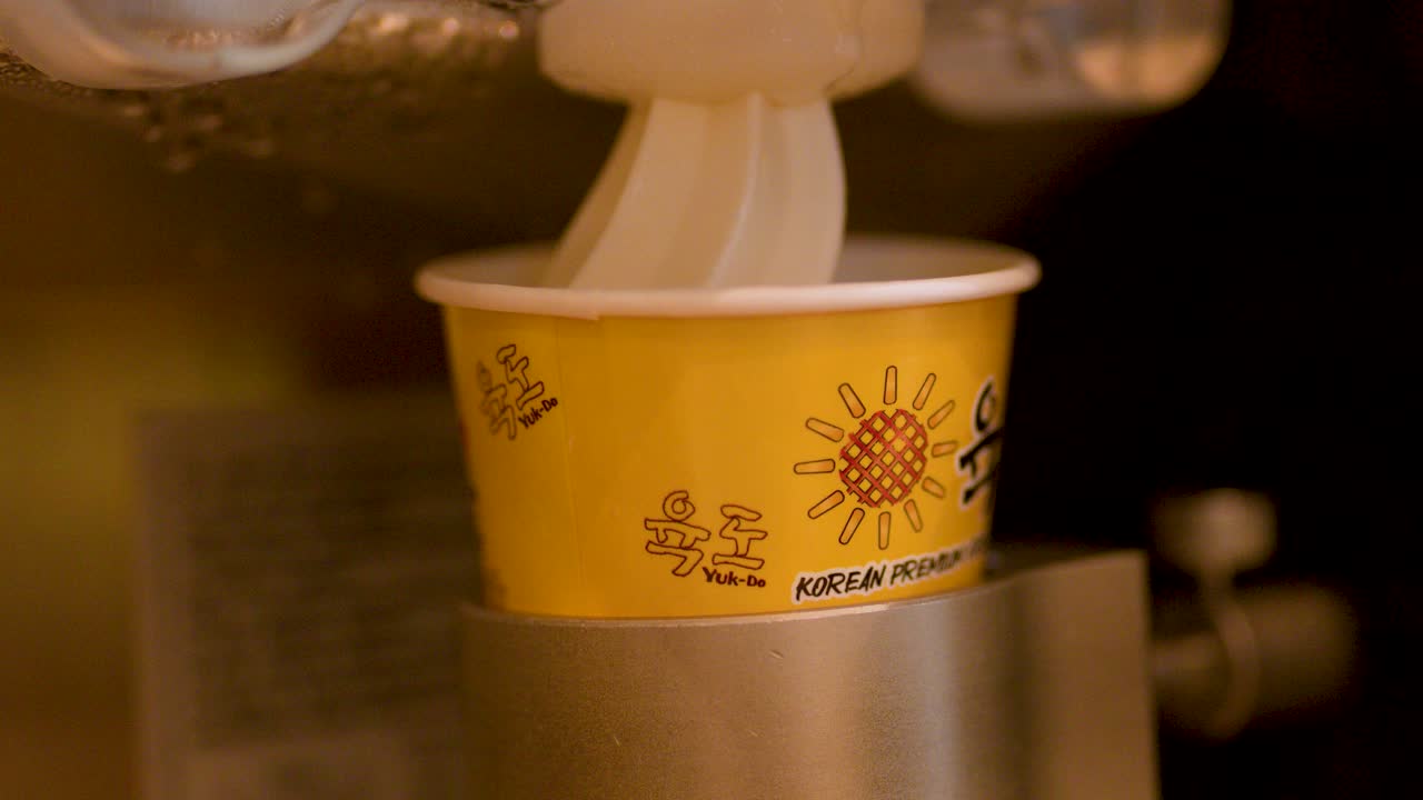 Soft serve ice cream dispensed into yellow cup under warm lights, hand retrieves finished dessert