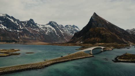 driving two colorful big cars over a bridge in beautiful lofoten islands in northern norway surrounded by mountains peaks, ocean, beaches and snow on a stormy and windy day with some sun