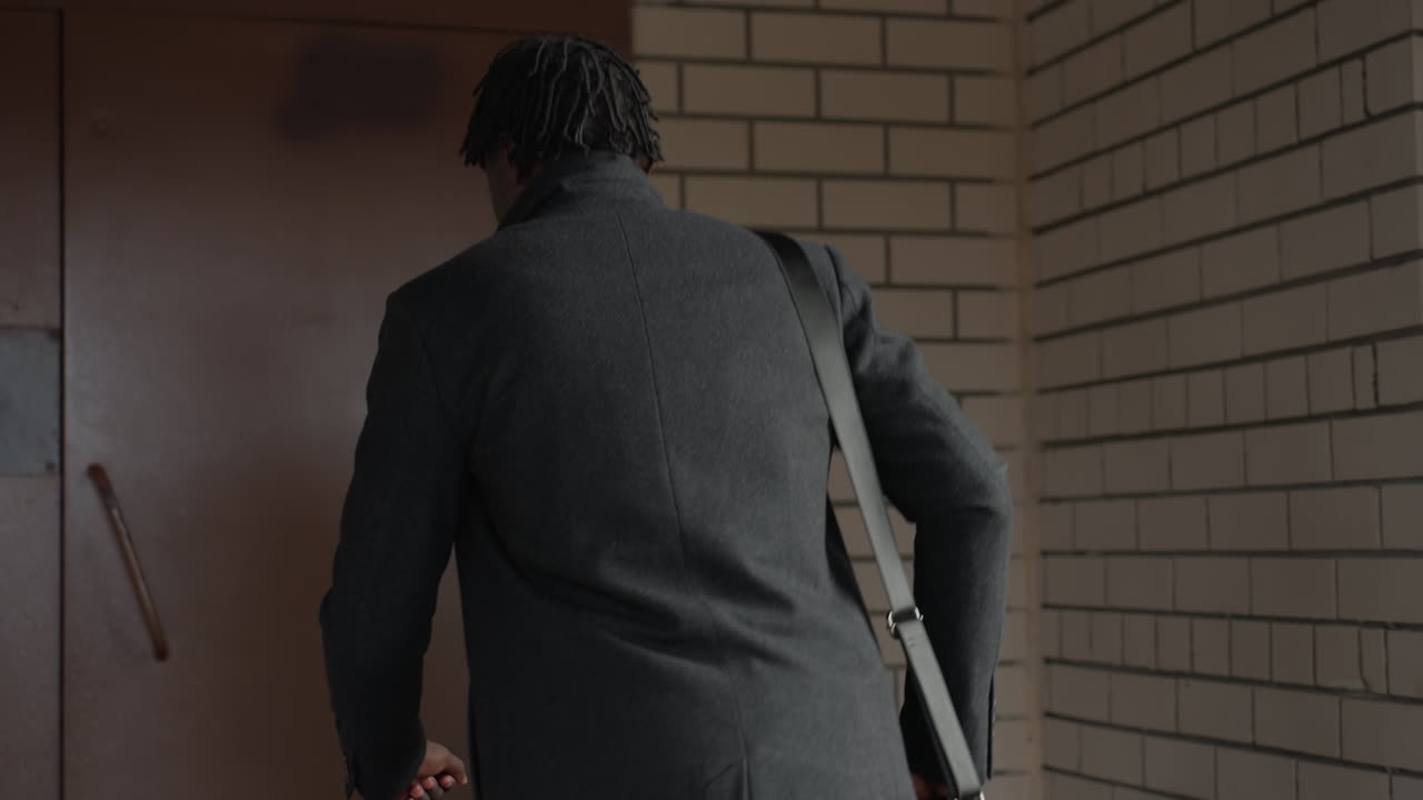 뒷모습 시점에서 다크 울 코트와 크로스바디 스트랩을 착용하고 벽돌 복도를 따라 스마트 출입구 쪽으로 걸어가는 스타일리시한 드레드락 남성이 보이네요. 금속 난간과 창틀, 시원한 조명 속에서 꾸준히 걷고 있어요.